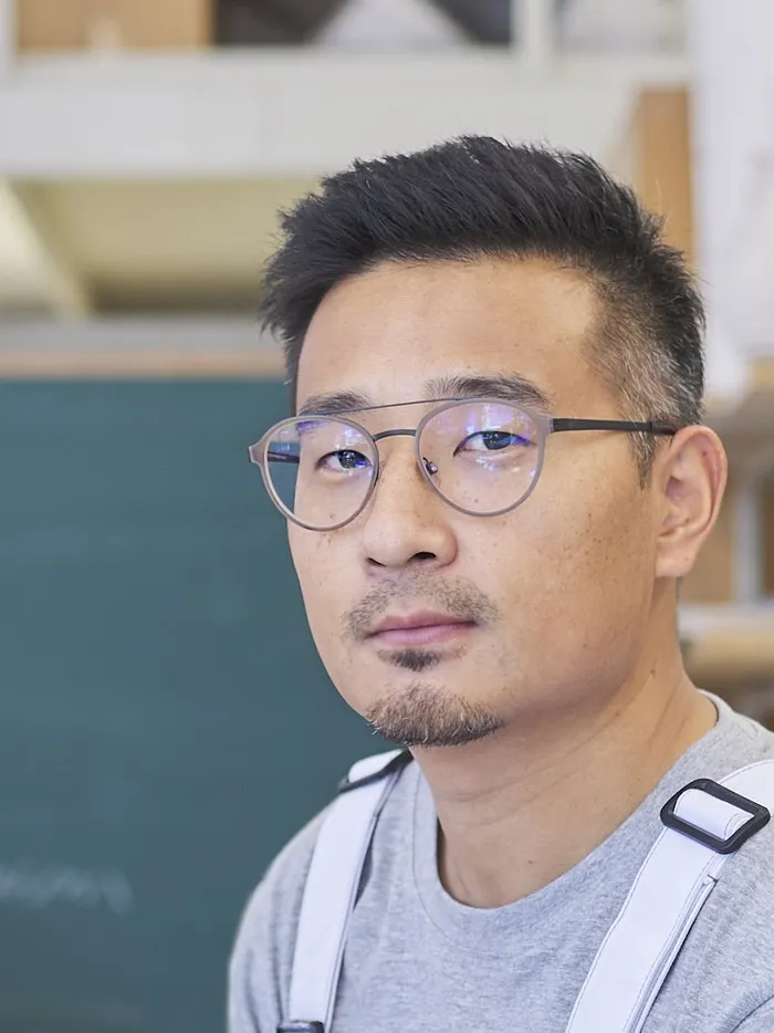 Porträt eines Mannes mit Brille in einer Werkstattumgebung, im Hintergrund eine grüne Tafel und Regale.