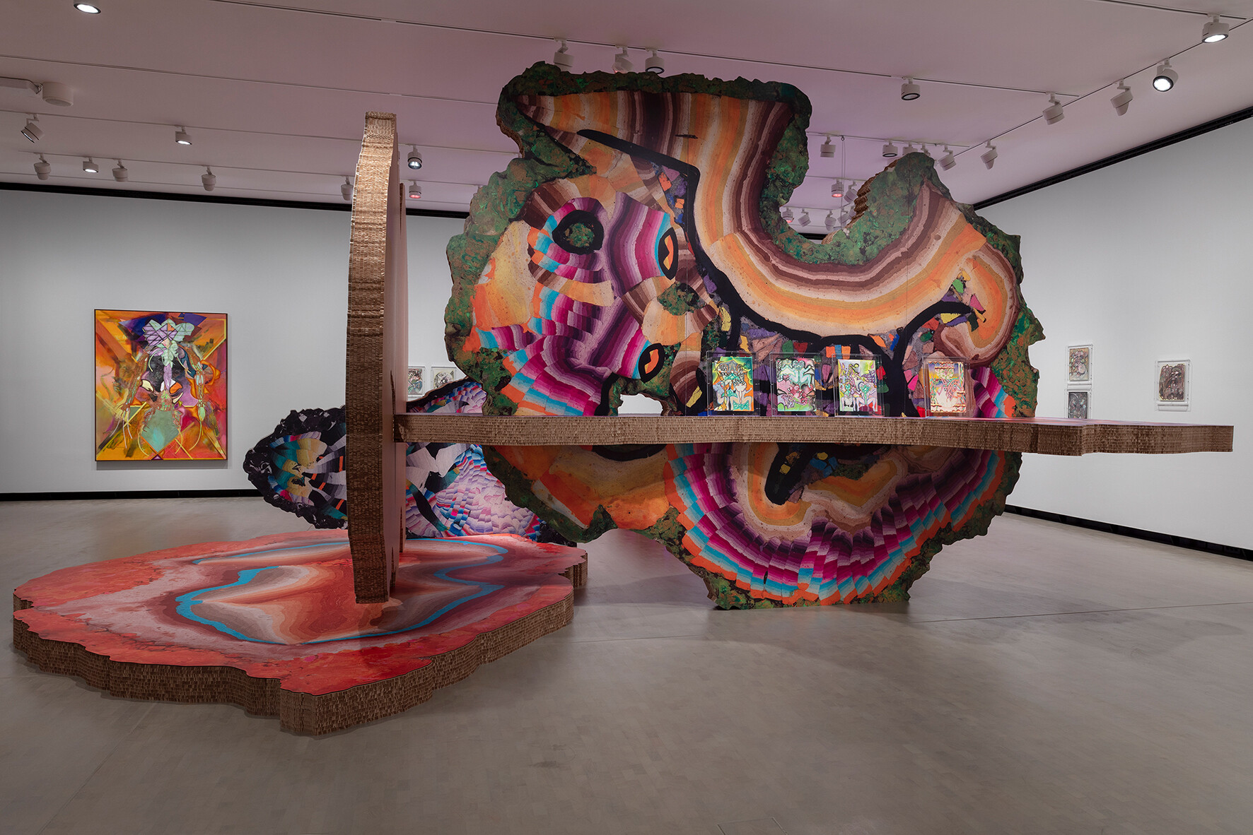 Großformatige Museumsinstallation in einem weißen Ausstellungsraum: Eine freistehende, organisch geformte, farbintensive Skulptur aus geschichteten, wellenförmigen Mustern in Pink-, Orange-, Grün- und Blautönen trägt mehrere kleine gerahmte Bilder. Ein langer horizontaler Steg durchschneidet die Form und verbindet sie mit einer zweiten Plattform am Boden, deren Oberfläche wie eine farbige Landschaft wirkt. An den Wänden hängen weitere Gemälde, Deckenstrahler beleuchten die Szene.