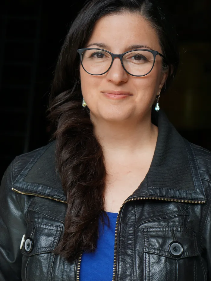 Portrait of a woman with dark hair worn to one side and black-framed glasses. She wears earrings, a blue top, and a black leather jacket. The background is dark and softly blurred, placing her face in focus.