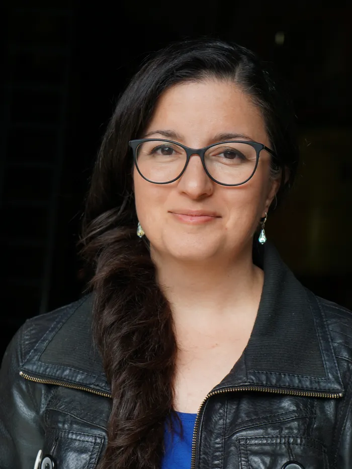 Porträt einer Frau mit dunklen, seitlich getragenen Haaren und schwarzer Brille. Sie trägt Ohrringe, ein blaues Oberteil und eine schwarze Lederjacke. Der Hintergrund ist dunkel und unscharf, wodurch ihr Gesicht im Fokus steht.