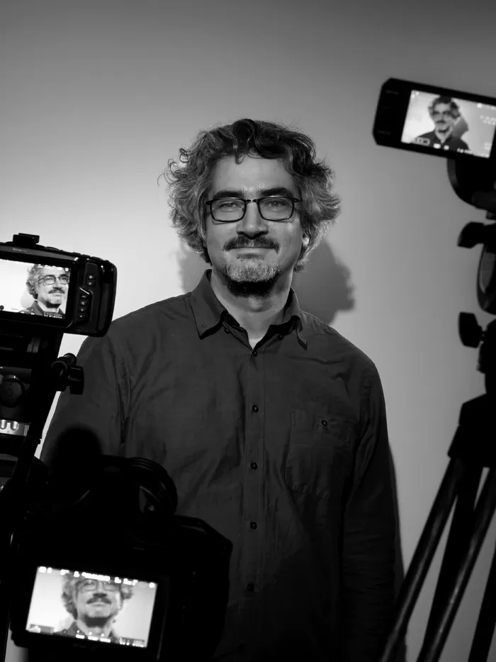 Schwarz-weiß-Porträt eines Mannes mit lockigem Haar und Brille, umgeben von Kameras auf Stativen.