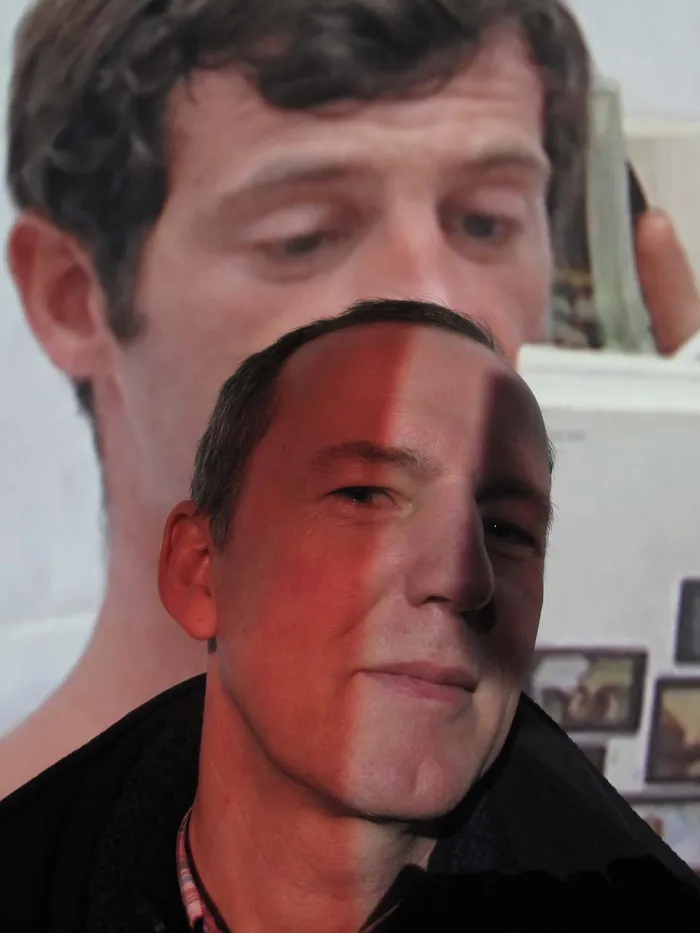 Porträt eines Mannes, dessen Gesicht teilweise von einer Projektion überlagert wird, im Hintergrund ein großformatiges Bild.