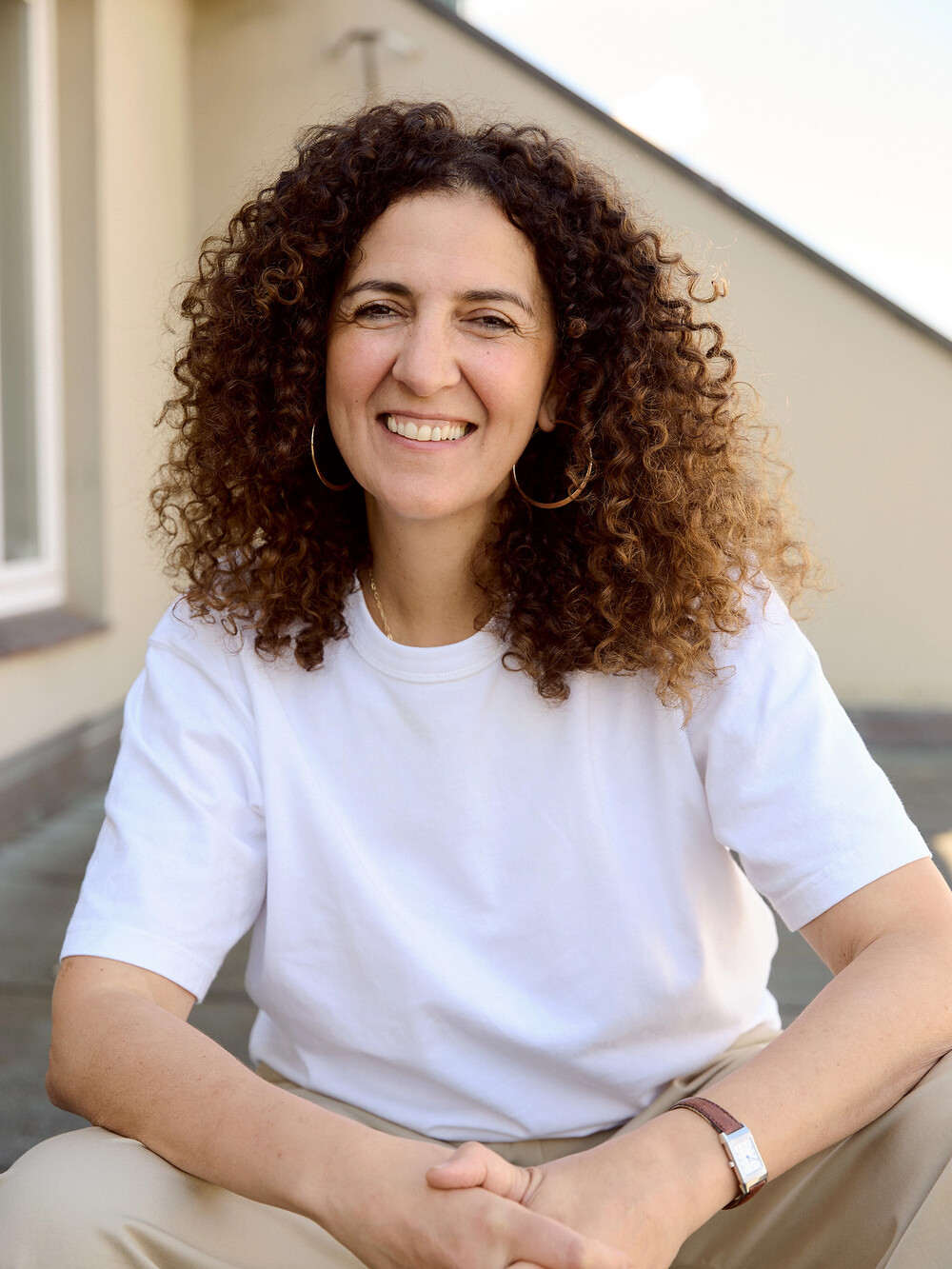 A smiling woman with long curly brown hair sits outdoors on steps. She is wearing a white T-shirt, beige pants, and large hoop earrings.