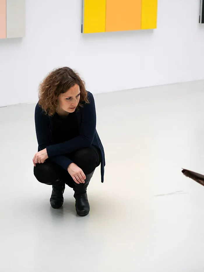 Woman crouching in an exhibition space with white walls, abstract color panels visible in the background.