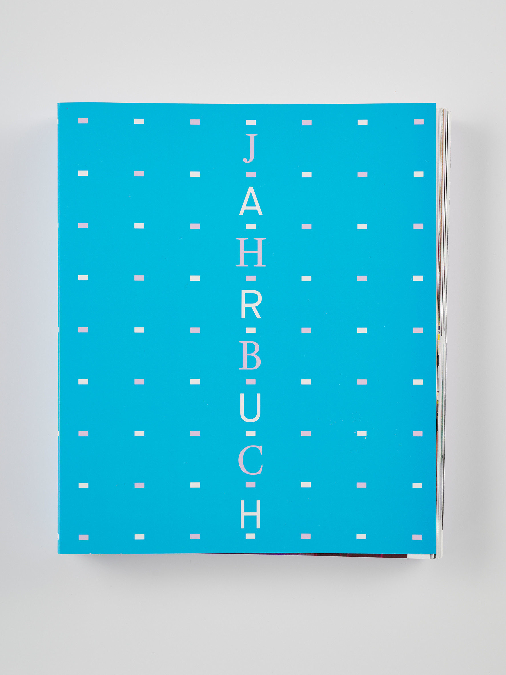 Türkisfarbenes Buchcover mit einem regelmäßigen Muster aus kleinen rechteckigen Punkten. In der Mitte steht der Titel „JAHRBUCH“ in vertikal gesetzten, hellen Buchstaben. Das Buch liegt auf einem neutralen Hintergrund.