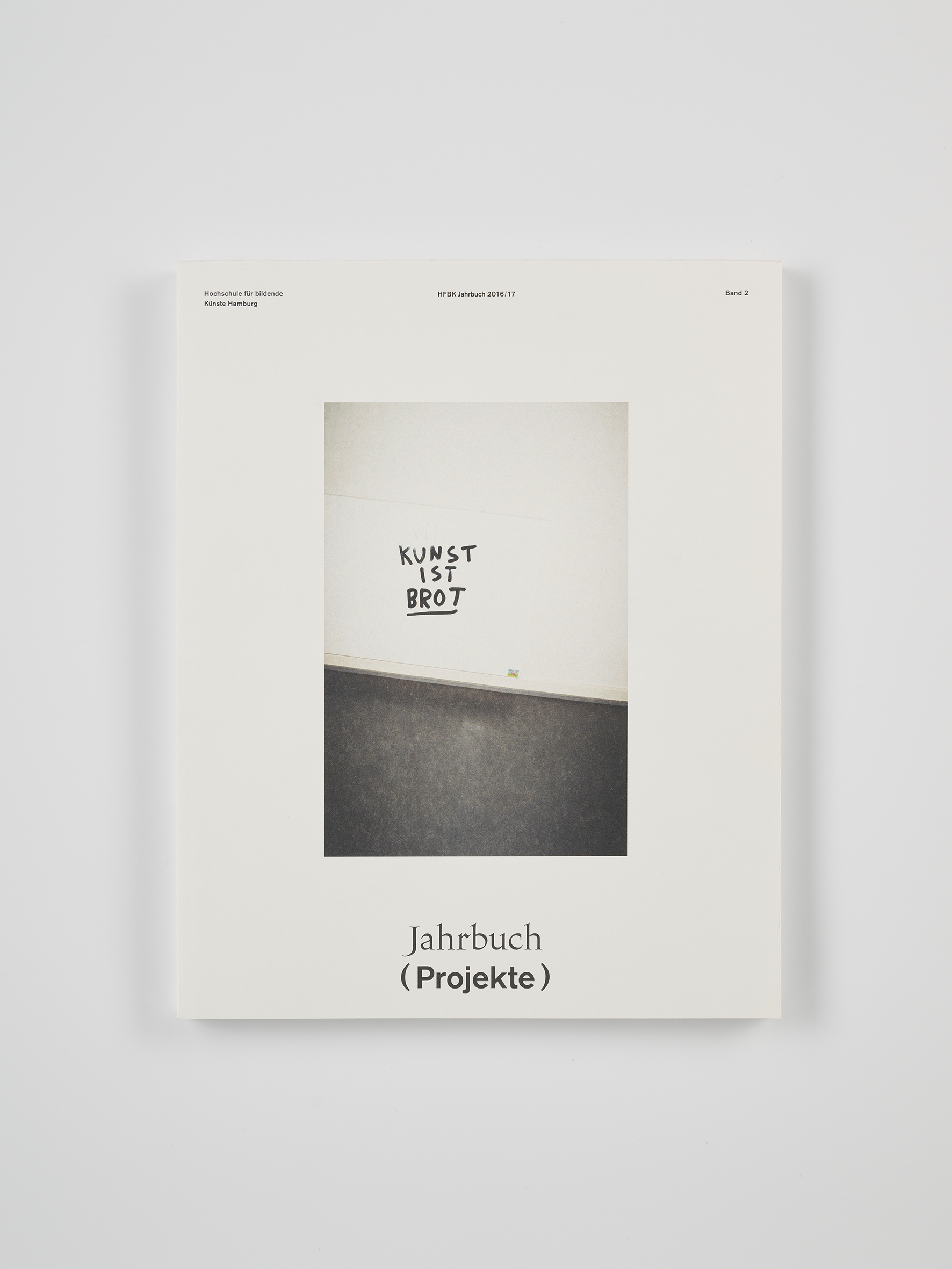 Weißes Buchcover mit einem zentrierten Foto einer Wand, auf der der Schriftzug „KUNST IST BROT“ steht. Darüber kleine Angaben zur HFBK Hamburg und zum Jahrbuch 2016/17, unten der Titel „Jahrbuch (Projekte)“. Das Buch liegt auf einem neutralen Hintergrund.