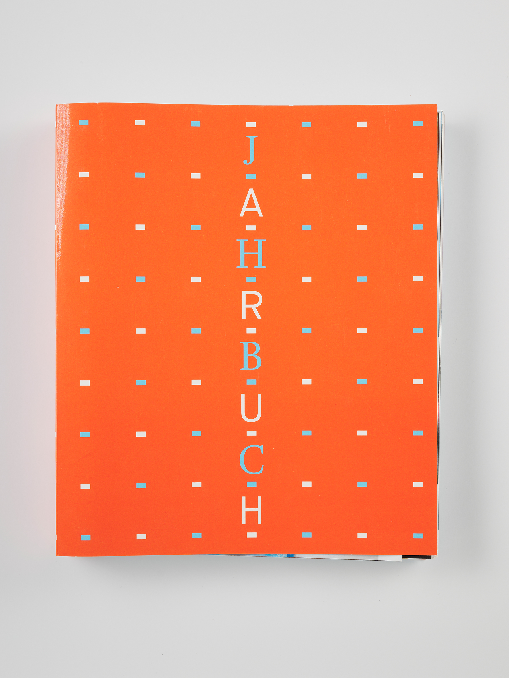 Orangefarbenes Buchcover mit einem regelmäßigen Muster aus kleinen rechteckigen Punkten. In der Mitte steht der Titel „JAHRBUCH“ in vertikal gesetzten, hellen Buchstaben. Das Buch liegt auf einem neutralen Hintergrund.