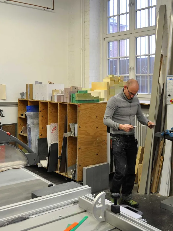 Man working in a workshop among machines, shelves, and material samples, holding a long workpiece.