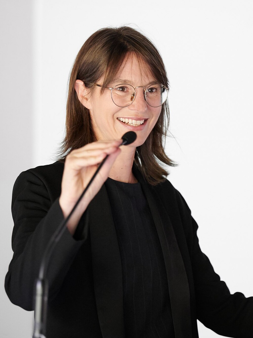 Frau mit Brille spricht lächelnd an einem Rednerpult in ein Mikrofon, vor hellem Hintergrund.