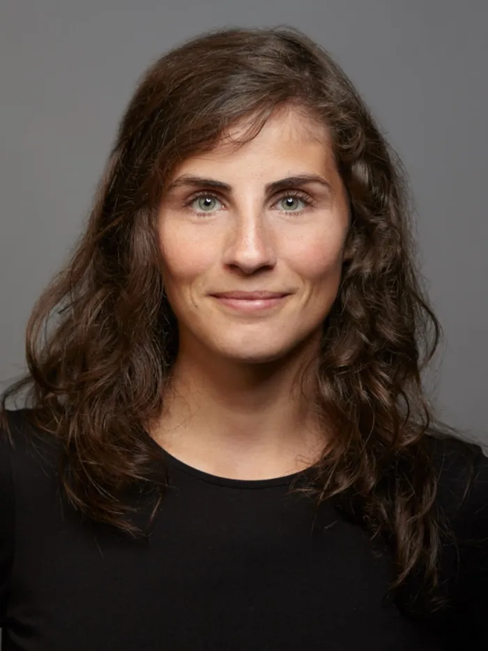 Porträt einer Frau mit lockigem Haar, neutraler Gesichtsausdruck, vor grauem Hintergrund.