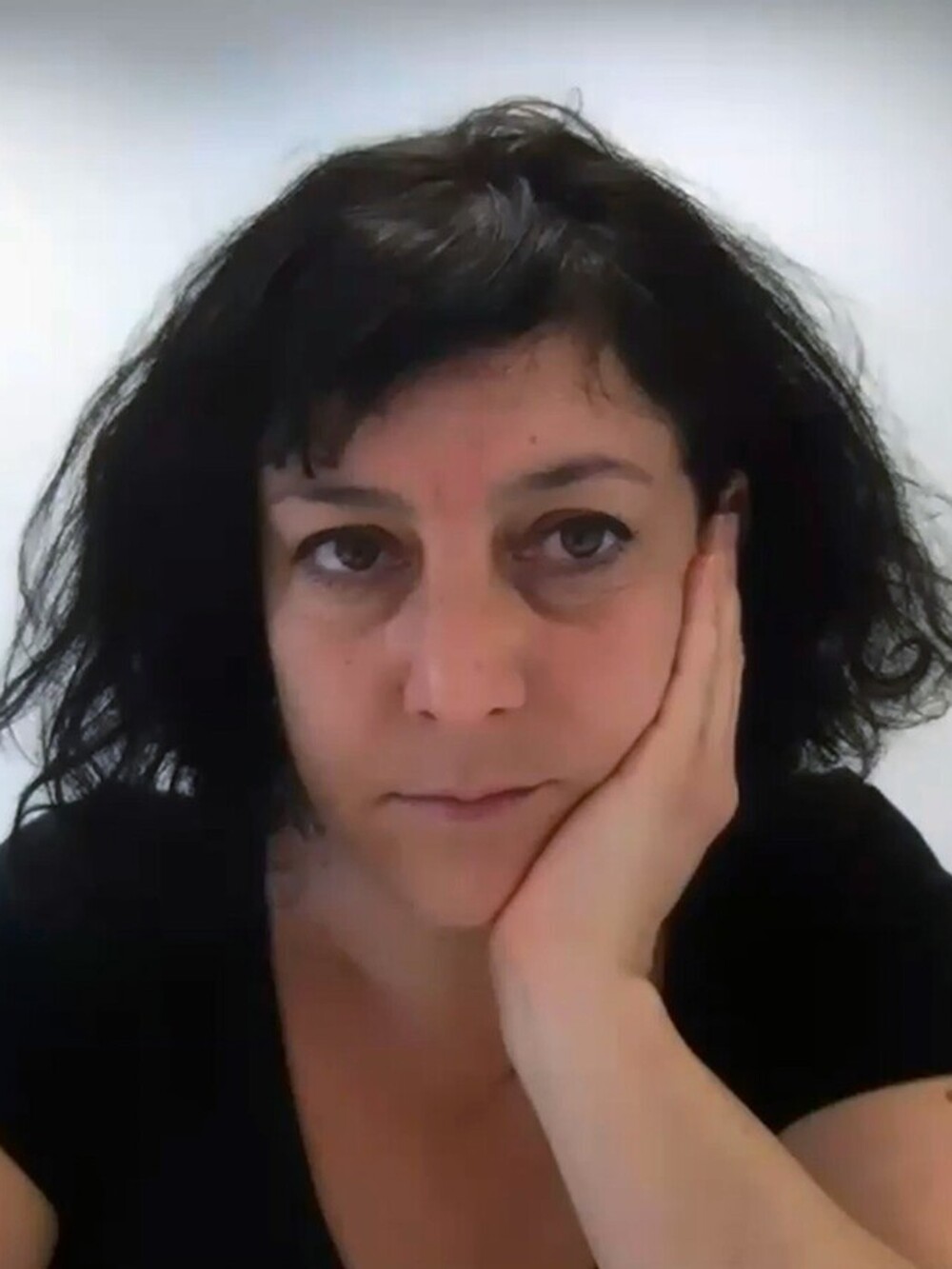 Eine Frau mit kurzen dunklen Haaren in einem schwarzen T-Shirt stützt ihren Kopf auf ihre Hand und blickt in die Kamera.