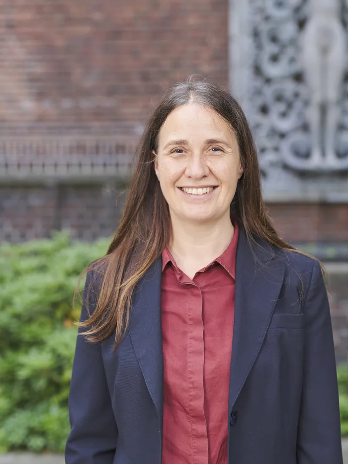 Portrait of a smiling woman with long hair standing outdoors in front of a brick wall and greenery.