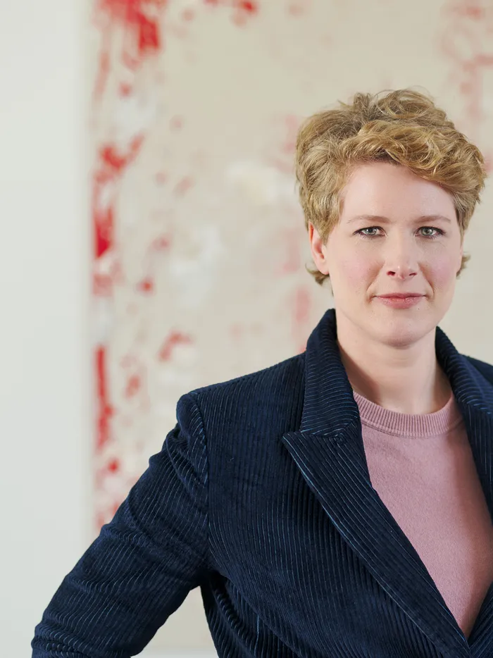 Porträt einer Frau mit kurzen blonden Haaren, die eine dunkle Jacke über einem rosafarbenen Oberteil trägt und vor einem großen abstrakten Gemälde mit roten und weißen Farbspritzern steht.