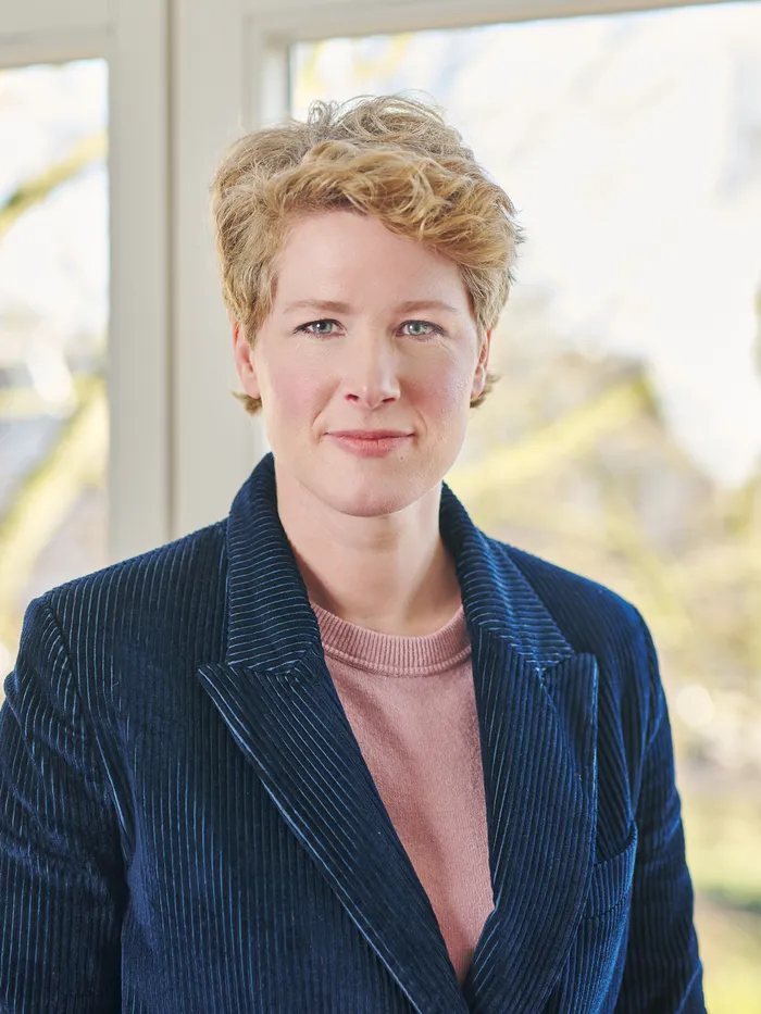 Portrait of a woman with short blonde hair wearing a dark jacket over a pink top, standing in front of a large abstract painting with red and white splashes.