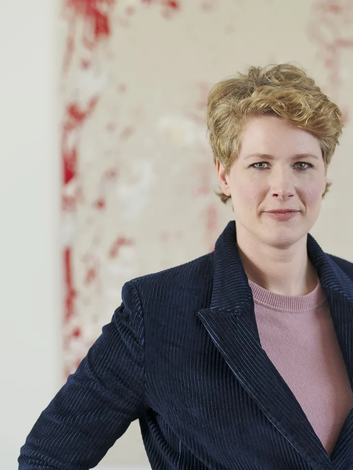 Porträt einer Frau mit kurzen blonden Haaren, die eine dunkle Jacke über einem rosafarbenen Oberteil trägt und vor einem großen abstrakten Gemälde mit roten und weißen Farbspritzern steht.