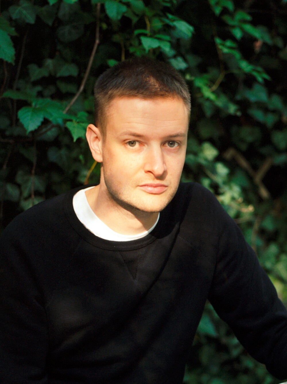 Portrait of a man with short hair sitting outdoors in front of dense foliage, wearing a dark top.