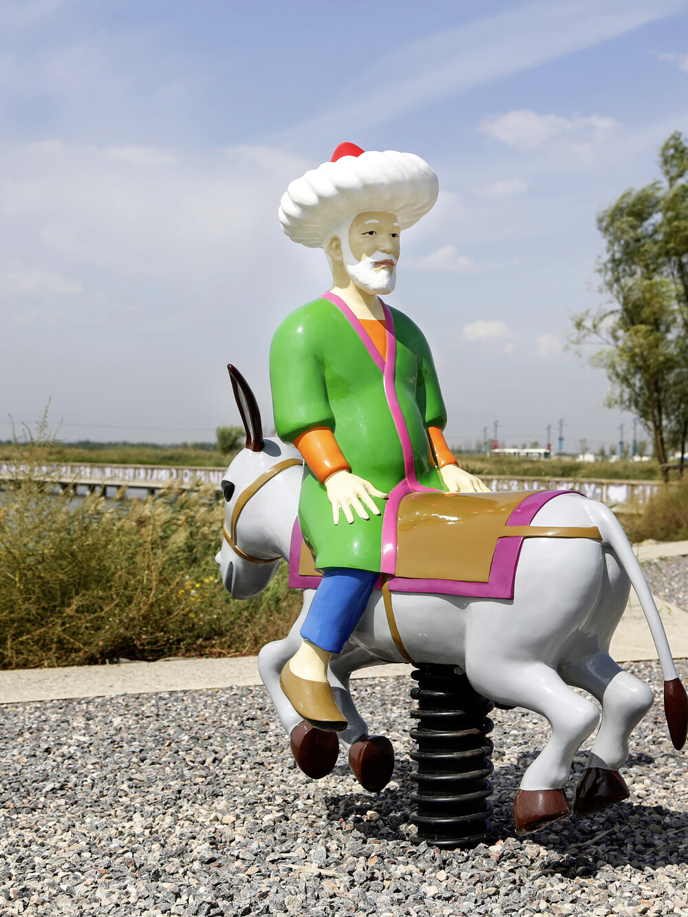 Auf einem Schotterfeld an einem Gewässer ist ein Wipptier installiert, das einen Esel und einen falsch herum auf dem Esel sitzenden Mann in grüner Kutte und mit Turban darstellt.