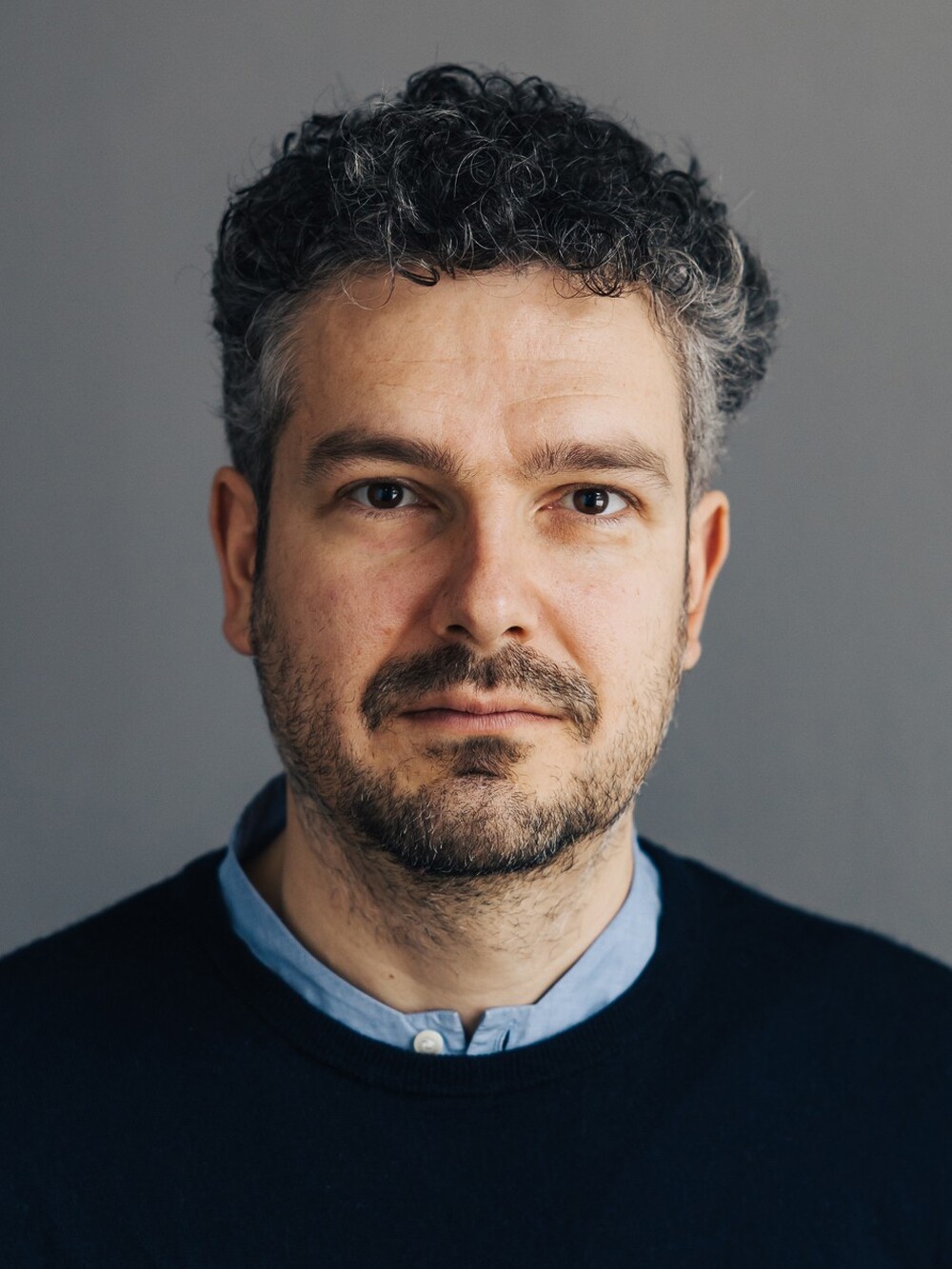 Ein Mann blickt in die Kamera. Er hat kurze schwarze, etwas lockigere Haare und einen Dreitagebart und trägt einen schwarzen Pulli.