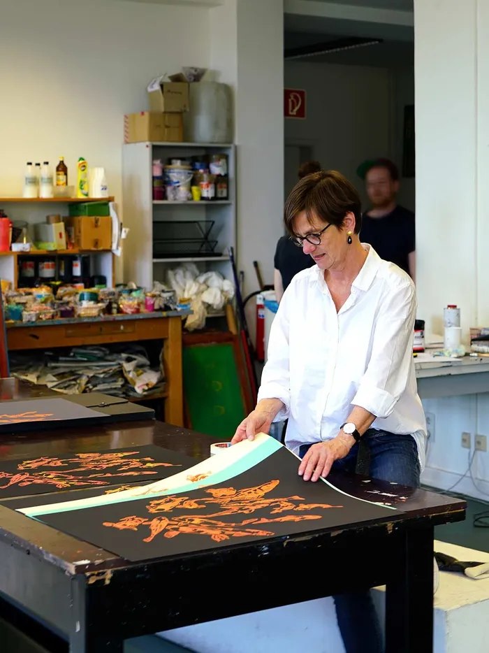 Woman working at a large table in a screen-printing workshop, aligning printed sheets of paper, surrounded by paints and tools.