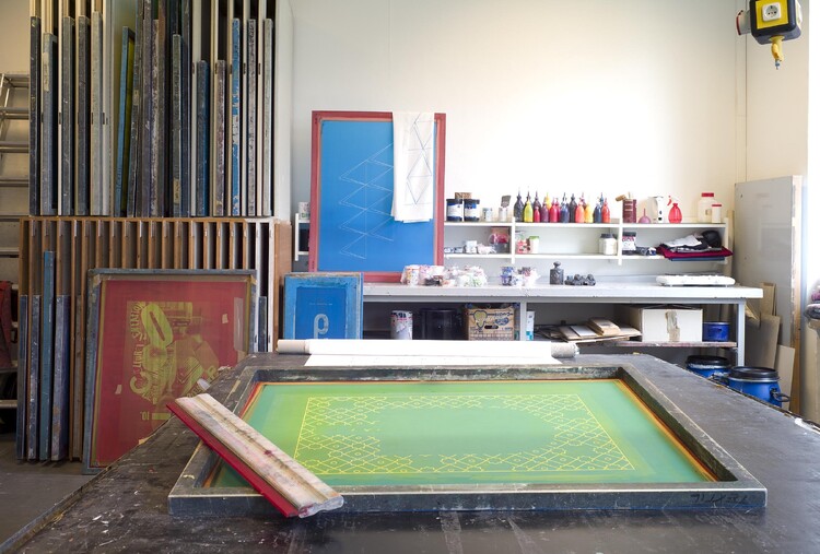 Ein beschichtetes Sieb mit einem Rakel im Vordergrund, dahinter Farb-Tuben und weitere Siebe