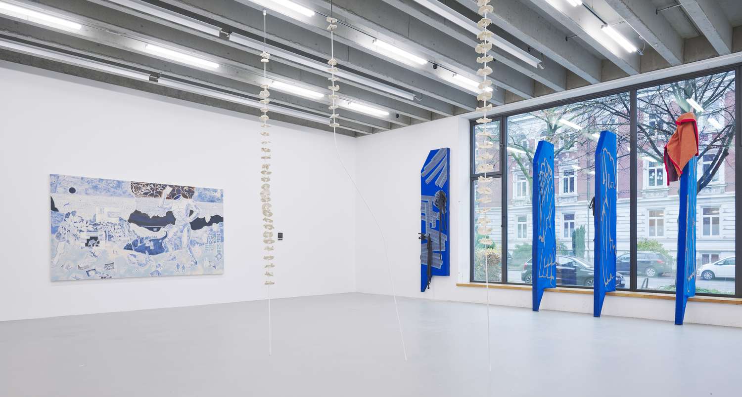Es ist ein heller Ausstellungsraum mit großen Fenstern zu sehen, in dem ein großformatiges Bild an der Wand hängt. In den Fenstern wurde eine große skulpturale Arbeit in Forma von vier großen blauen Fahnen angebracht. Der Raum hat einen grauen Boden.