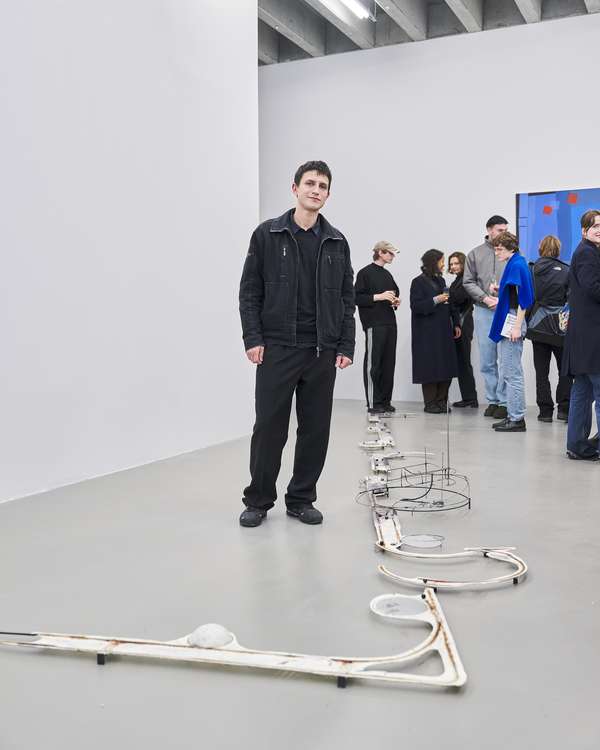 Ein junger Mann, schwarz gekleidet, steht in einem weißen Ausstellungsraum und schaut freundlich in die Kamera. Am Boden befindet sich seine Installation aus schwarzen Draht und anderen Materialien. Im Hintergrund sind einige Besucher*innen der Ausstellung zu sehen.